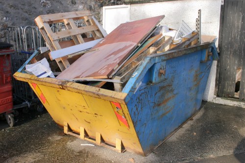 A large, rectangular skip with a yellow front panel and blue sides is positioned on a paved driveway in an outdoor area. The skip is filled with various types of waste materials, including wooden pallets, panels, and planks which are stacked haphazardly, with some leaning against the sides. The wooden components exhibit different textures, some smooth and painted, others rough and weathered, with colors ranging from natural wood tones to white and reddish-brown. The skip's metal surface shows signs of rust and wear, particularly on the blue sections. In the background, a white wall, a wooden fence, and parts of other waste management equipment are partially visible, with natural daylight illuminating the scene. This setting reflects a typical object handled by Hounslow House Clearance services in the local area for rubbish disposal and clearance in the Hounslow region.
