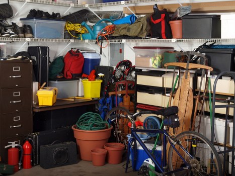 The image shows a cluttered garage interior with a variety of household and outdoor items. In the foreground, there are several plastic flower pots of different sizes, some stacked and others placed individually, with textures ranging from smooth to slightly rough. Nearby, a blue bicycle with a black seat and handlebars is partially visible, leaning against wooden shelves that hold an assortment of storage boxes, including black, grey, and clear plastic containers. A green garden hose is coiled around itself in a terracotta pot, situated next to additional empty pots on the floor. To the left, a black filing cabinet with five drawers stands next to a portable radio or speaker, and a yellow plastic container is perched on top of a small black work surface or table. Above, metal wire shelves are filled with various items such as folded fabric, a red bag, a cardboard box, and loose cables, including a yellow extension cord and a tangle of electrical wires. The overall scene depicts a typical cluttered storage space used for household and garden equipment, consistent with rubbish removal services in Hounslow or nearby areas.
