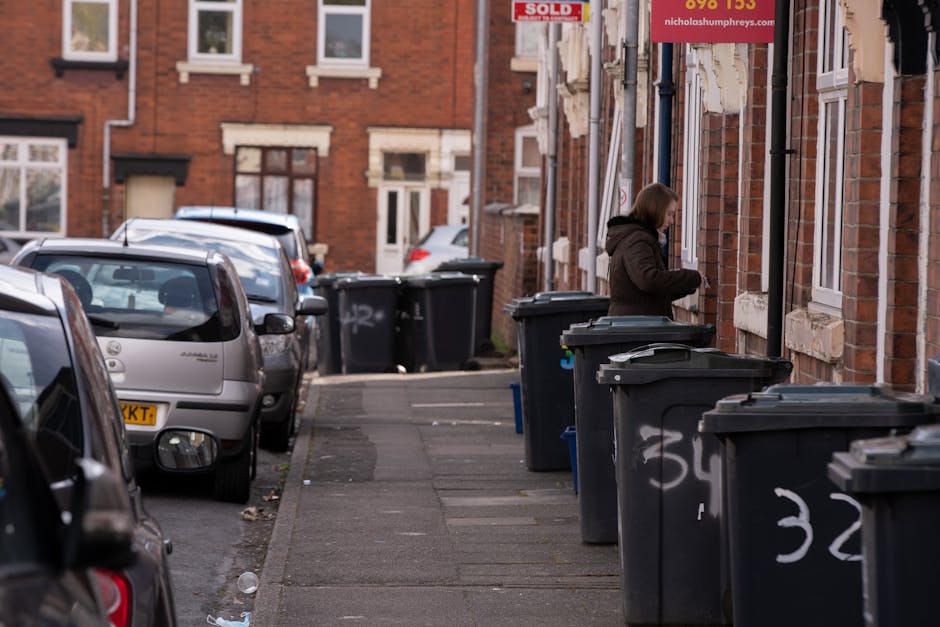 In a city street scene, a large pile of mixed waste is accumulated around three overflowing rubbish bins. The waste includes cardboard boxes, plastic bags, paper, and discarded packaging, some of whic