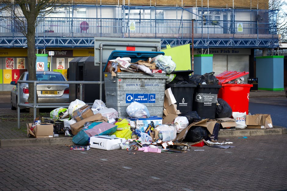 A large collection of mixed waste and rubbish bags, cardboard boxes, and loose paper cluttering a section of paved sidewalk in front of a commercial building. The waste includes black plastic bags, fl